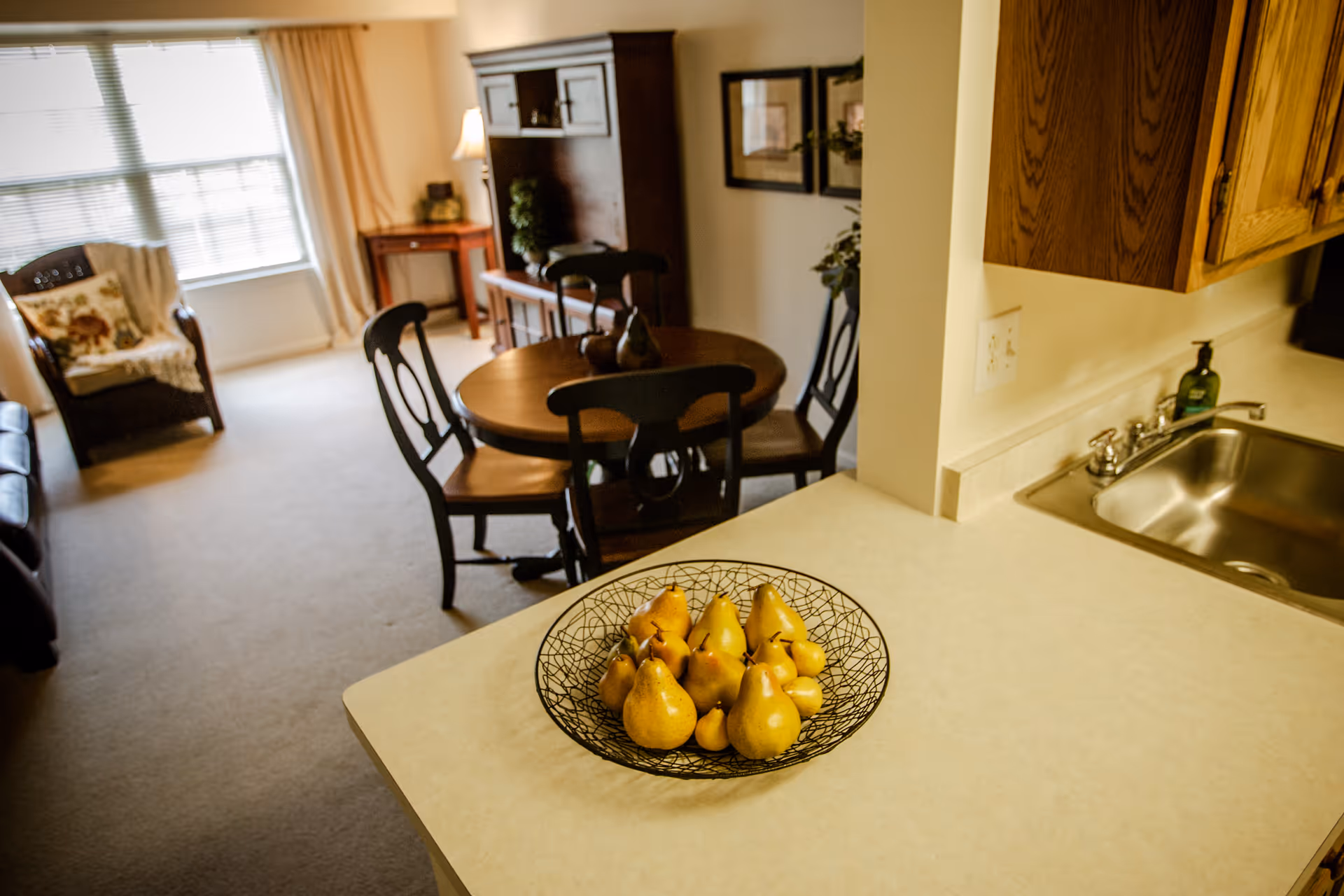 View of a cozy living and dining area from a kitchen counter. The counter has a decorative bowl filled with yellow pears. In the background, there is a round wooden dining table with four chairs, a wooden cabinet, a small side table with a lamp, and a cushioned armchair near a window with blinds and curtains.