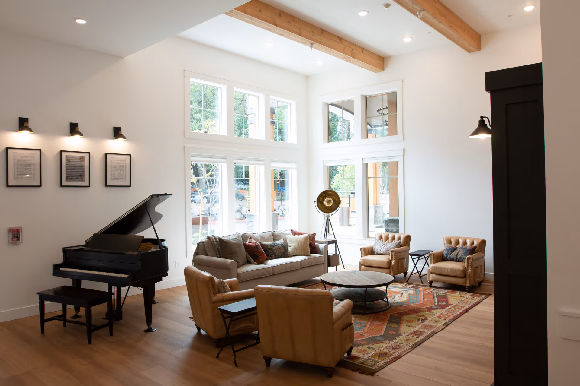A bright and spacious living room with large windows letting in natural light. The room features a beige sofa with multiple cushions, three tan leather armchairs, a round wooden coffee table on a colorful patterned rug, and a black grand piano with a matching bench. The walls are white with wooden beams on the ceiling and framed pictures on one wall. A vintage-style floor lamp stands near the windows.