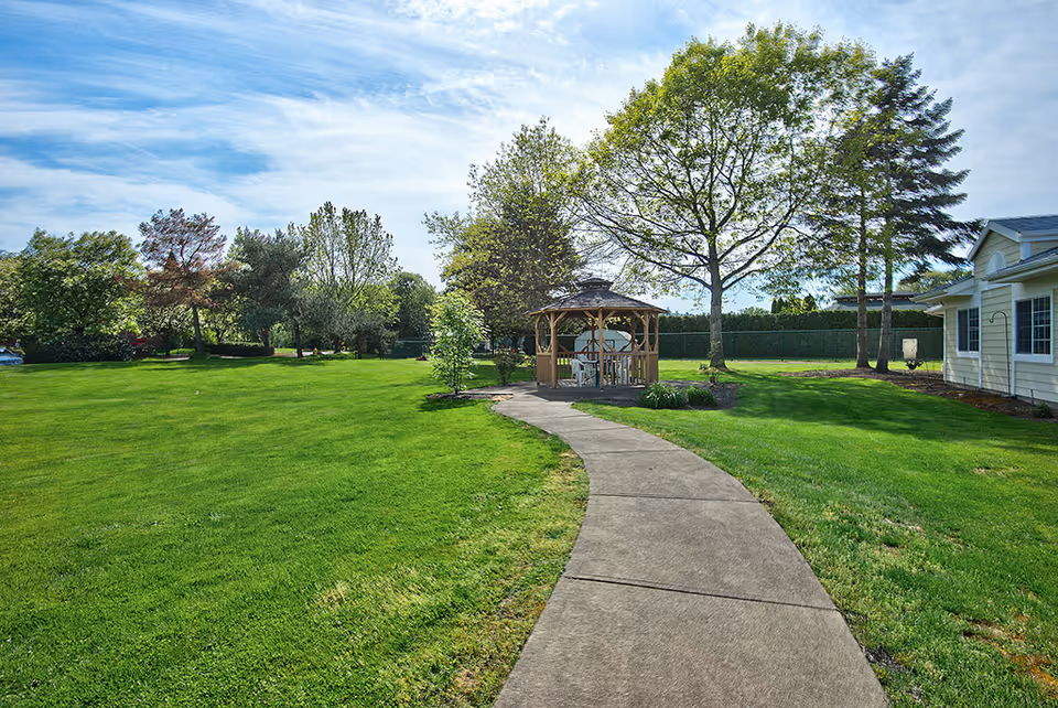 A paved walkway through a green lawn leads to a wooden gazebo near trees and a light-colored building.