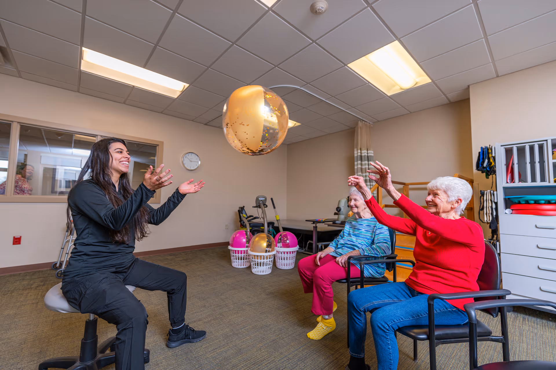A young woman and two elderly women sitting in chairs in a senior living community activity room, playing with a gold and transparent beach ball. The room has exercise equipment, baskets with balls, and storage shelves in the background.