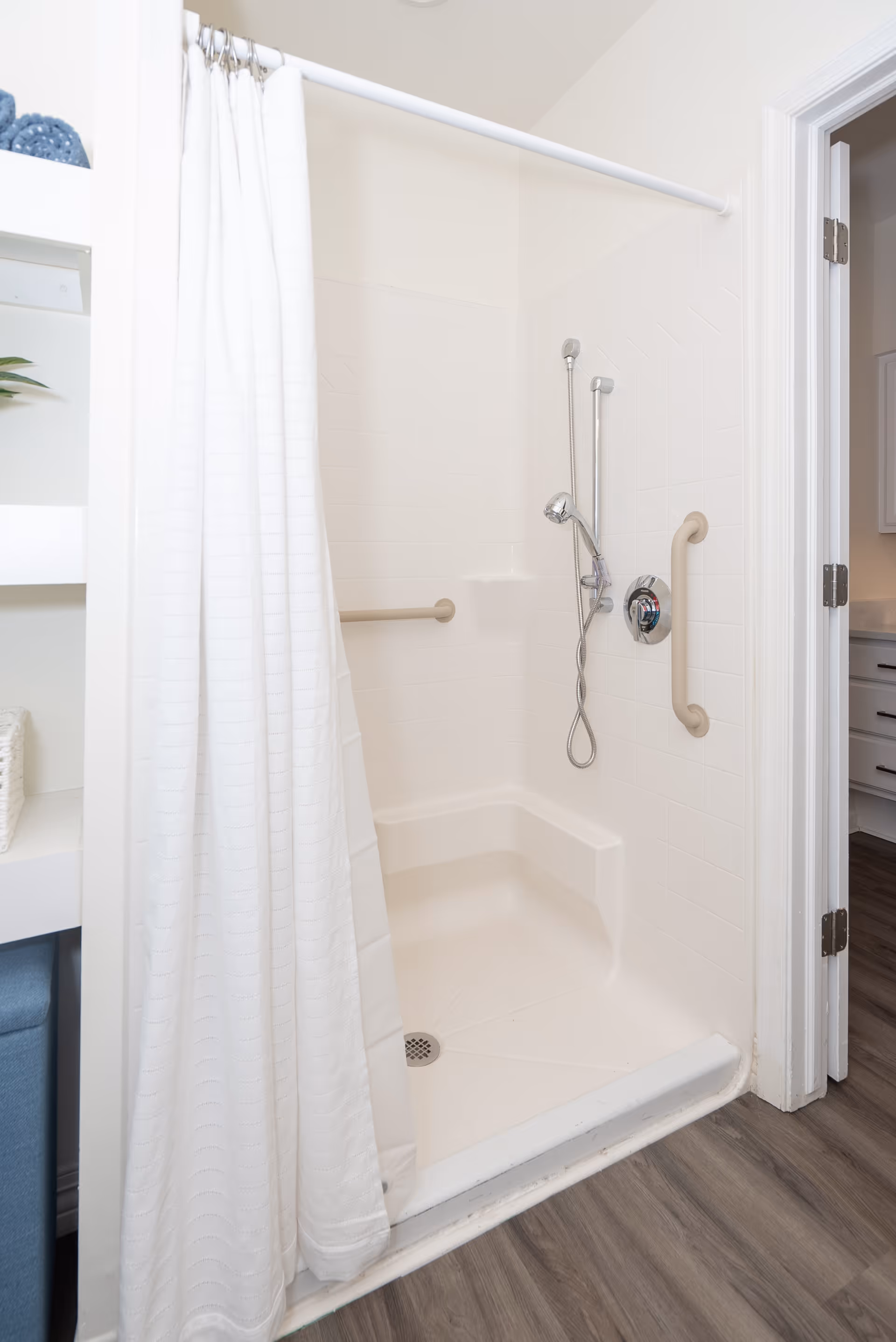 A clean, white walk-in shower with a built-in seat and grab bars on the walls. The shower has a handheld showerhead and a white shower curtain partially drawn to the side. The floor outside the shower is wood-style laminate, and a doorway to another room is visible on the right.