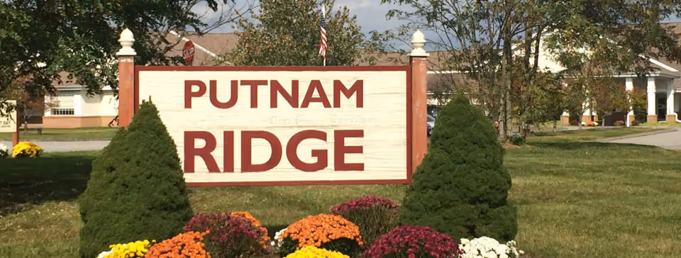 Outdoor view of a large sign reading 'PUTNAM RIDGE' surrounded by green shrubs and colorful flowers, with a building and trees in the background under a partly cloudy sky.
