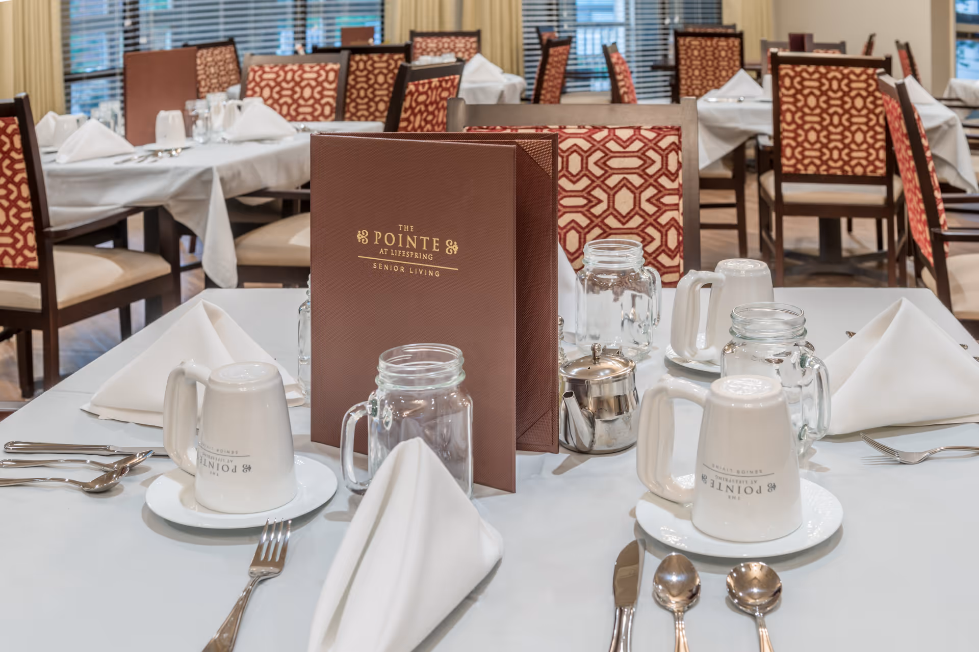 A dining table set for a meal in a senior living facility with white tablecloth, folded white napkins, silverware, glass mugs, and a brown menu card labeled 'The Pointe at Lifespring Senior Living'. The background shows multiple tables and chairs with patterned upholstery and large windows with blinds.