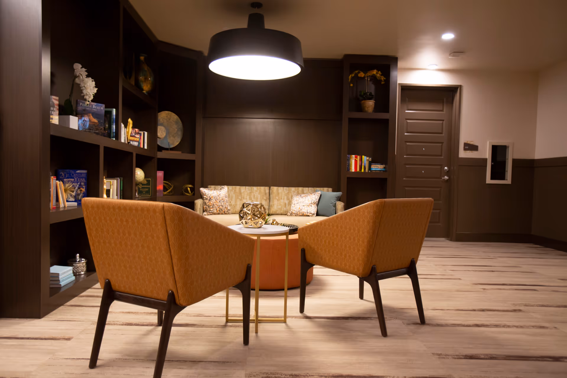 A cozy seating area in a senior living facility with two orange armchairs facing a round ottoman and a beige couch with decorative pillows. The space features built-in dark wood shelves filled with books and decorative items, a modern hanging light fixture, and a closed door in the background.