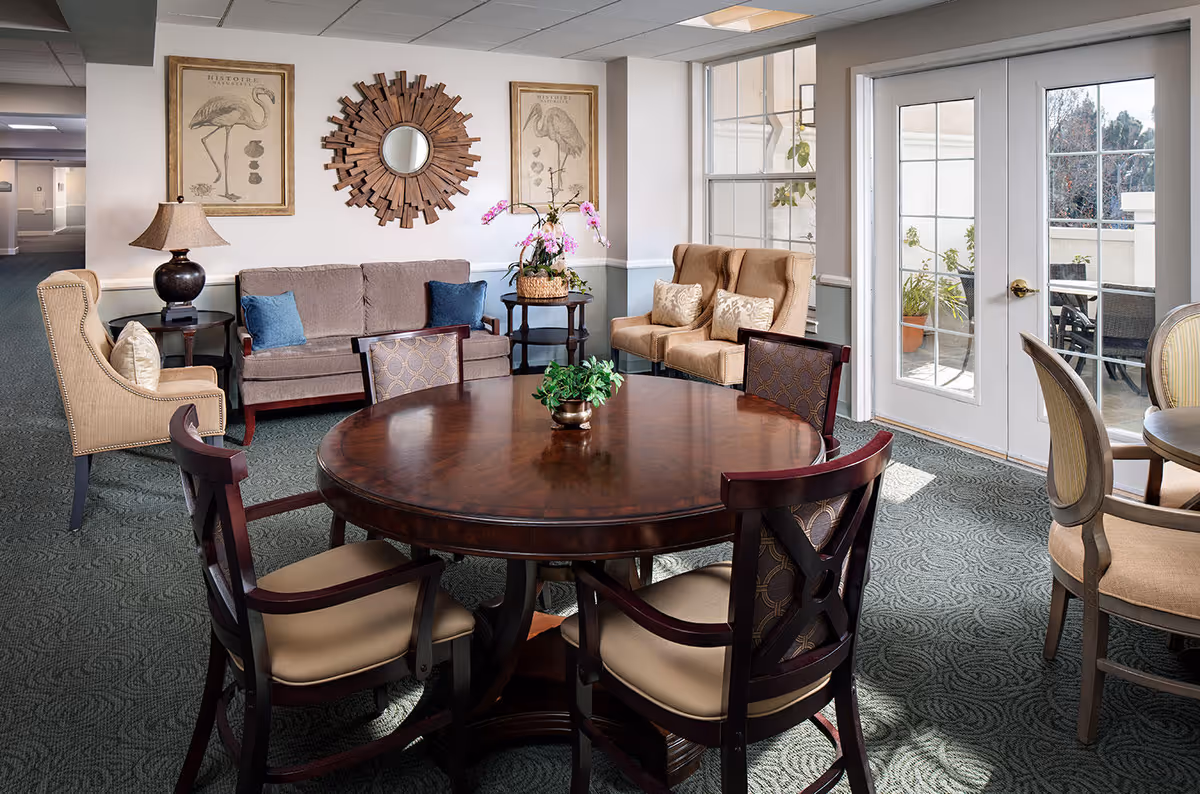 A cozy senior living common area with a round wooden table surrounded by four cushioned chairs in the foreground. Behind the table, there is a beige sofa with blue pillows, two beige armchairs with patterned cushions, and a side table with a lamp and a basket of flowers. The walls are decorated with two framed flamingo prints and a round wooden sunburst mirror. Large windows and a glass door let in natural light and provide a view of an outdoor patio with plants and seating.