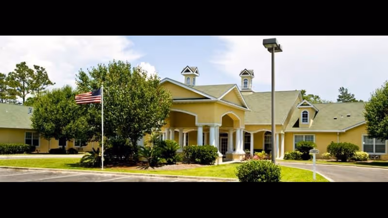 Exterior view of a single-story yellow building with a green roof, white columns at the entrance, surrounded by trees and bushes, with an American flag on a flagpole in front and a parking area visible.