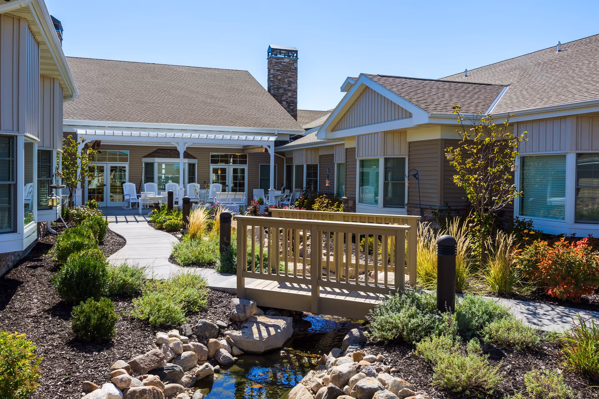 Outdoor courtyard area of a senior living facility with a small wooden bridge over a rock-lined stream, surrounded by landscaped bushes and plants. The building has beige siding, multiple windows, and a covered patio with white chairs under a clear blue sky.