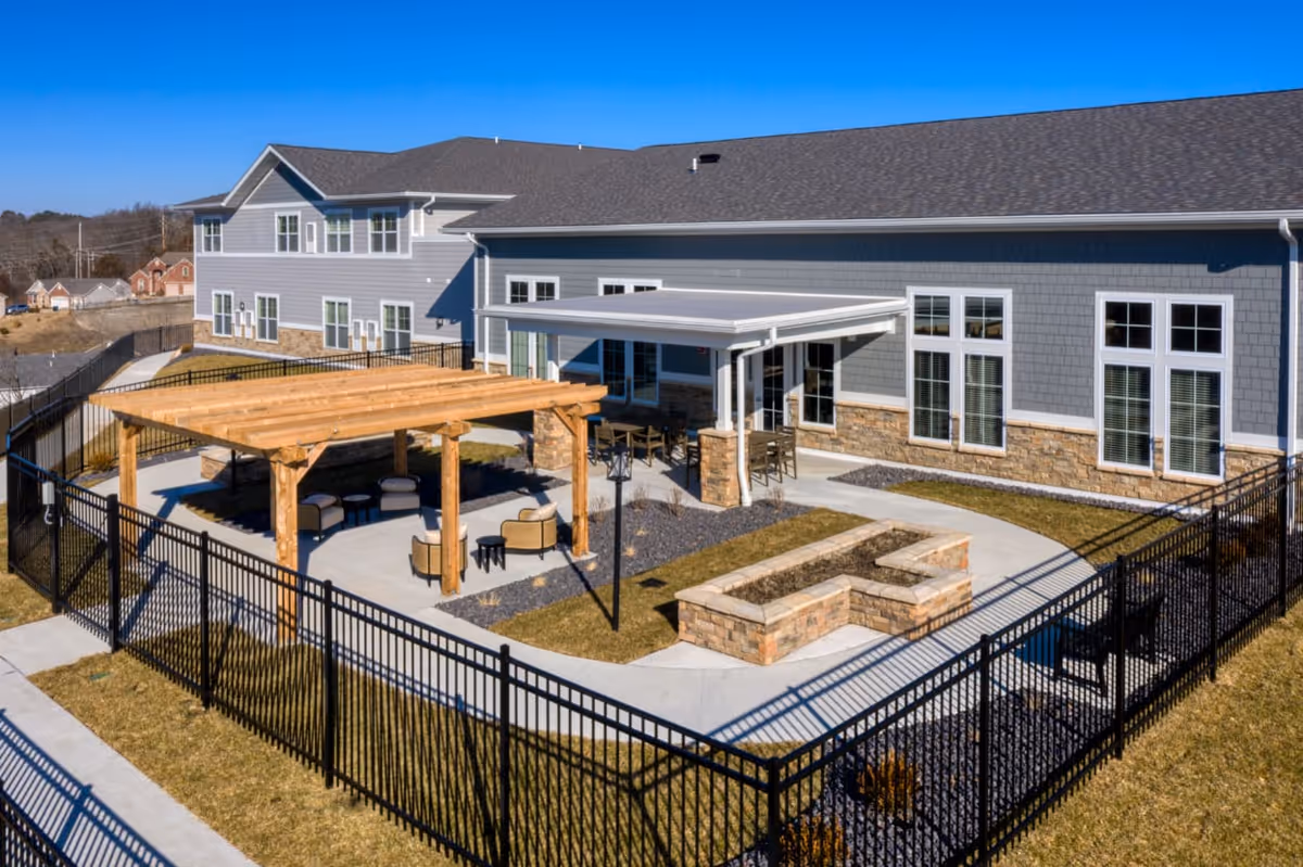 Outdoor patio area at Cedarhurst Senior Living of Arnold featuring a fenced-in space with a wooden pergola, seating arrangements including chairs and tables, a covered patio section attached to the building, and landscaped pathways with stone planters.