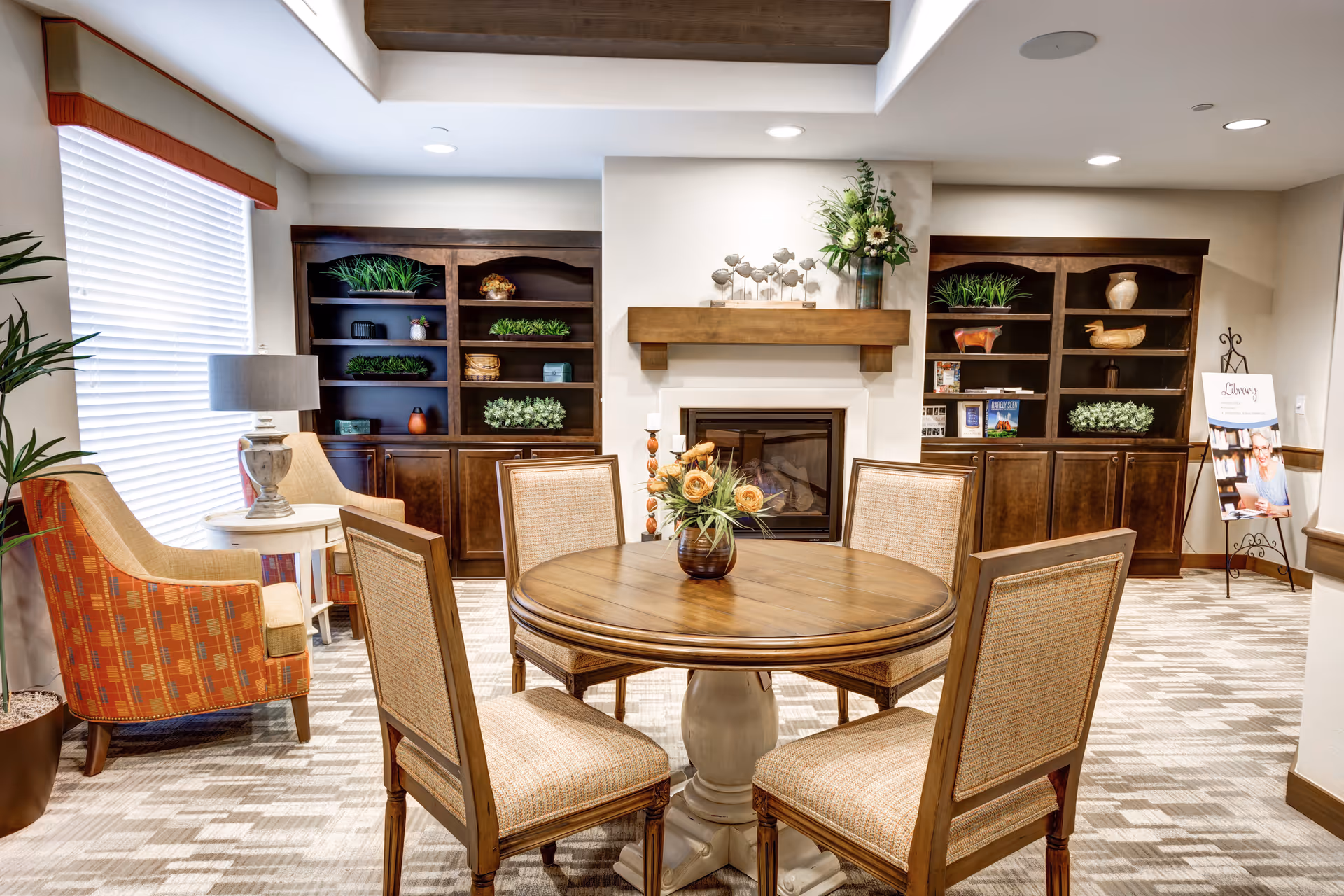 A cozy senior living common area with a round wooden table surrounded by four cushioned chairs. Behind the table is a fireplace with a wooden mantel decorated with a vase of flowers and small sculptures. On either side of the fireplace are dark wooden built-in shelves with various decorative items and greenery. To the left, there is a comfortable armchair next to a small side table with a lamp, and a large window with blinds and a valance. The room has a warm and inviting atmosphere with soft lighting and neutral tones.