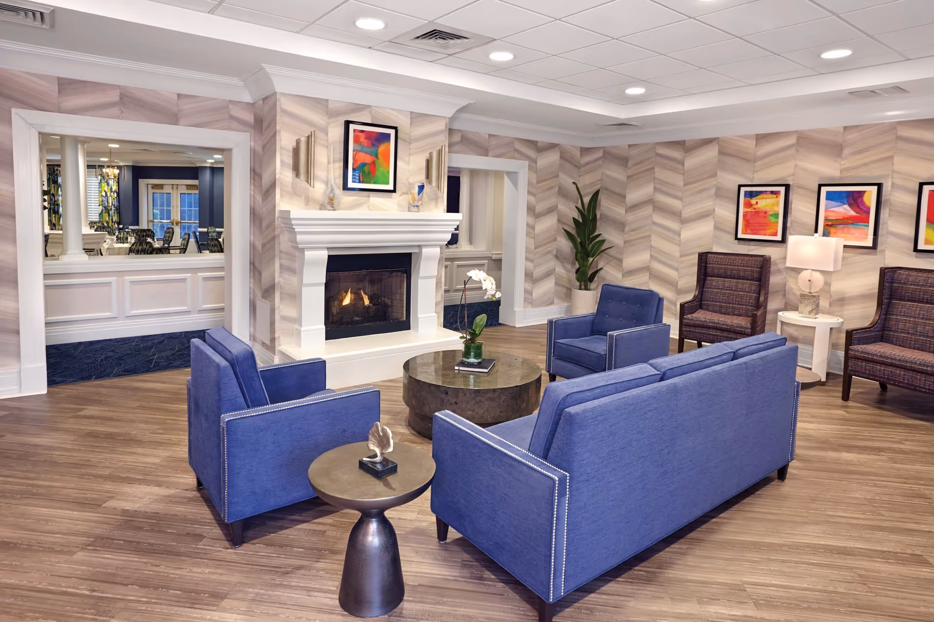 A cozy living room area in an assisted living facility featuring a blue sofa and two matching blue armchairs arranged around a round coffee table with a small plant. There is a white fireplace with a colorful abstract painting above it. The walls have a chevron pattern wallpaper, and additional seating includes two patterned armchairs with a white side table and lamp between them. Three colorful abstract paintings hang on the wall behind the chairs. A large plant is placed in the corner, and through an open doorway, a dining area with tables and chairs is visible.