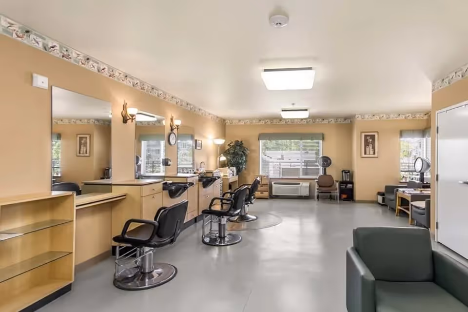 Bright interior salon with barber chairs, mirrors, styling stations, and seating in a senior living facility.