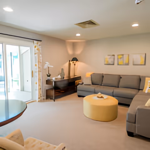 A cozy living room with a large gray sectional sofa, a round yellow ottoman in the center, a wooden side table with a lamp and decorative items, three abstract paintings on the wall, and sliding glass doors leading to an outdoor area.