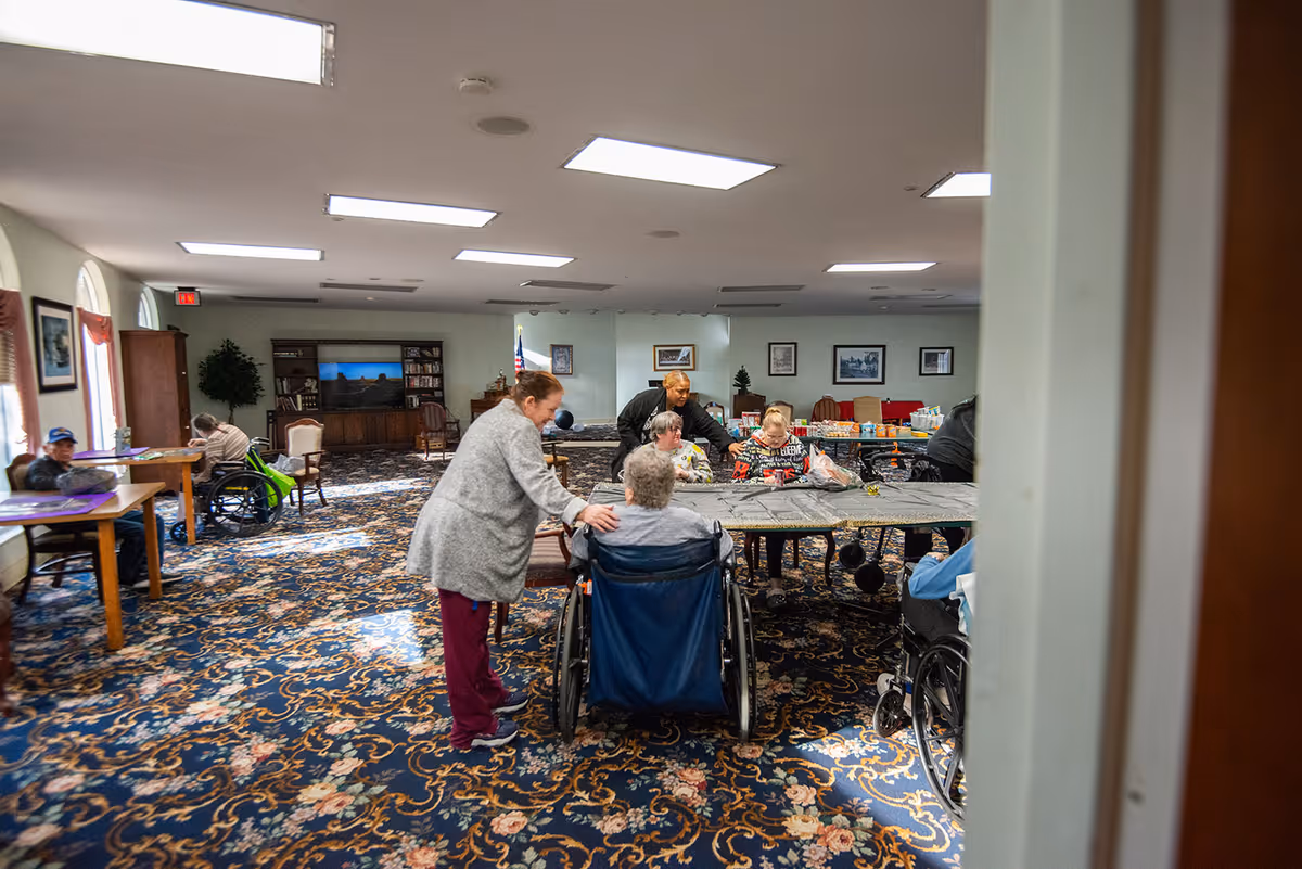 A group of elderly people, some in wheelchairs, gathered around tables in a spacious room with patterned carpet and bright overhead lighting. A caregiver is interacting with one of the residents. The room has large windows with curtains, framed pictures on the walls, and a television in the background.