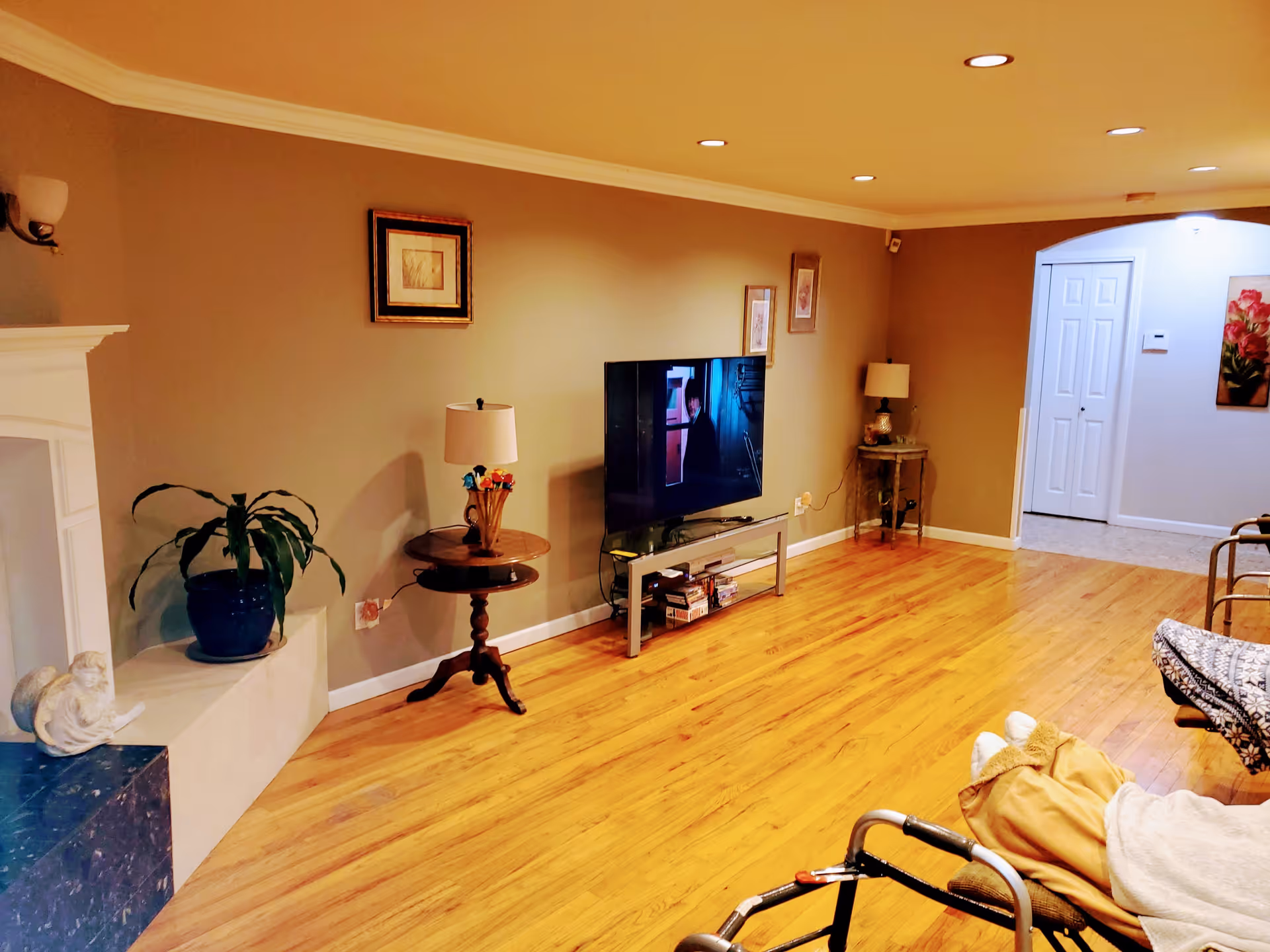 A cozy living room with wooden flooring, beige walls, and recessed ceiling lights. There is a flat-screen TV on a stand against the wall, flanked by two small tables with lamps. A potted plant sits on a ledge near a white fireplace. In the foreground, a person is seated in a wheelchair, covered with a blanket. The room leads to a hallway with white double doors and a floral painting on the wall.