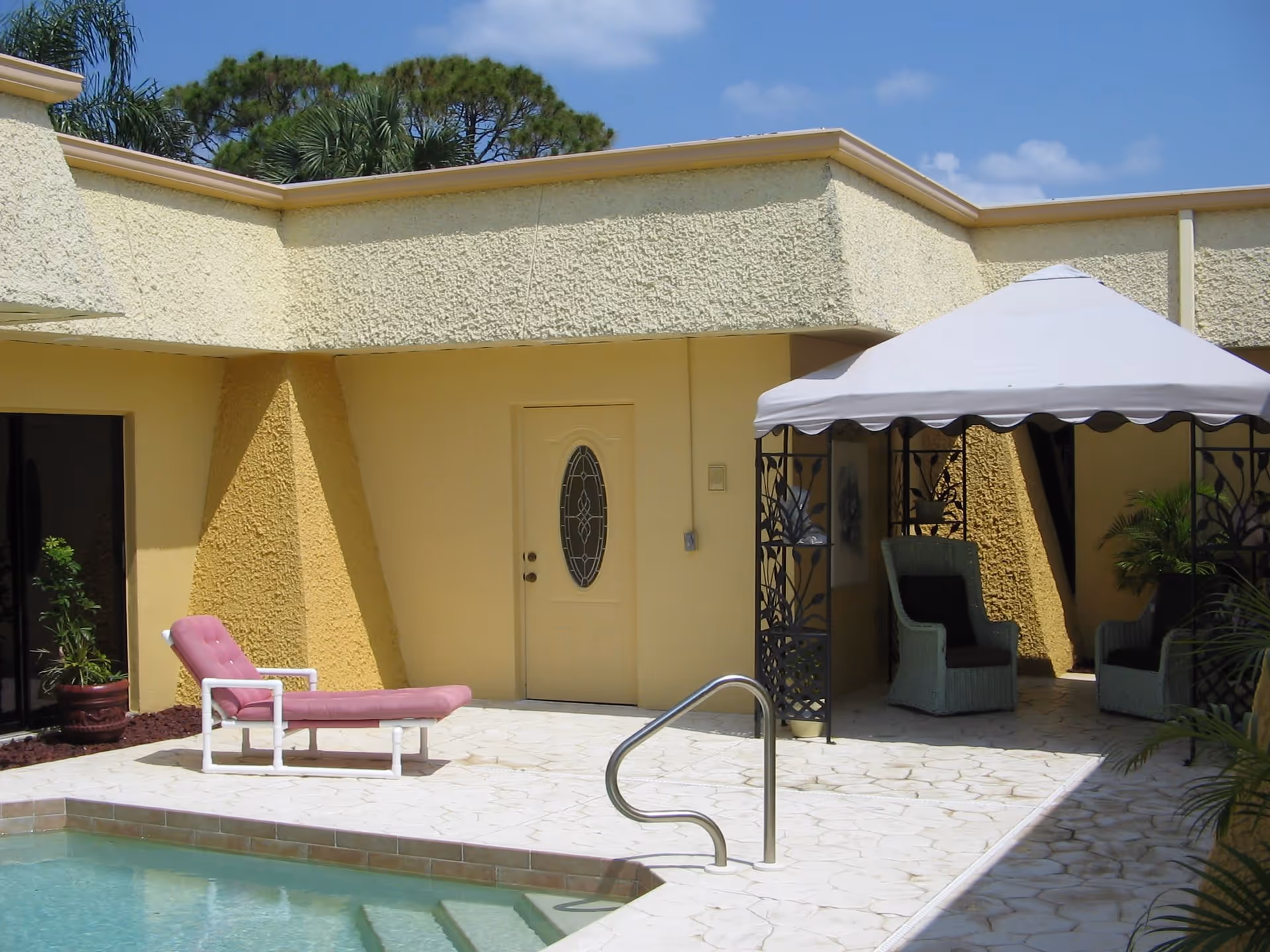 Outdoor patio area with a swimming pool, a pink cushioned lounge chair, a white canopy with two wicker chairs underneath, and yellow textured walls of the building in the background under a clear blue sky.