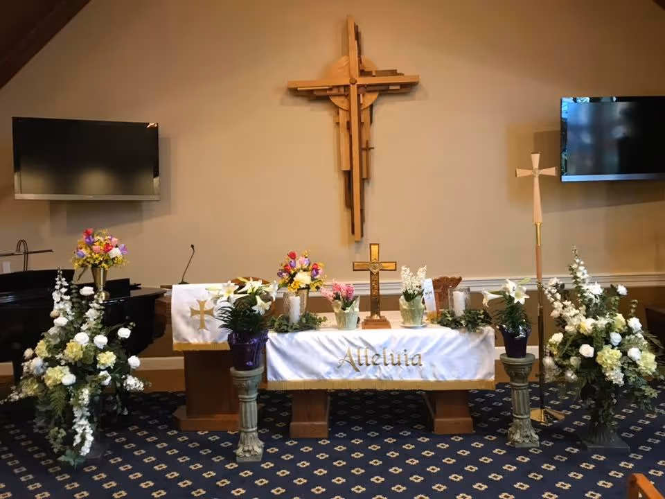 Interior chapel altar with a wooden cross on the wall, an 'Alleluia' altar cloth, floral arrangements and two wall-mounted TVs.