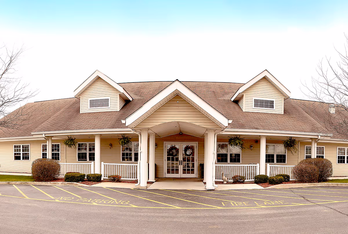 Front exterior view of a single-story building with beige siding and a brown shingled roof. The entrance features a covered porch with white columns and double glass doors decorated with wreaths. There are several windows along the front, hanging plants on the porch, and neatly trimmed bushes. The parking area in front has yellow painted markings indicating no parking and fire lane zones.