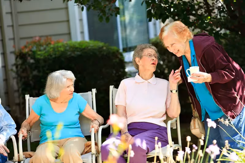 Three elderly women outdoors at a senior living facility. Two women are seated on white chairs, one wearing a turquoise shirt and beige pants, the other wearing a light pink shirt and purple pants. The third woman, standing and holding a white cup and saucer, is wearing a burgundy jacket over a blue shirt. They appear to be engaged in conversation in a garden or patio area with greenery and flowers around them.