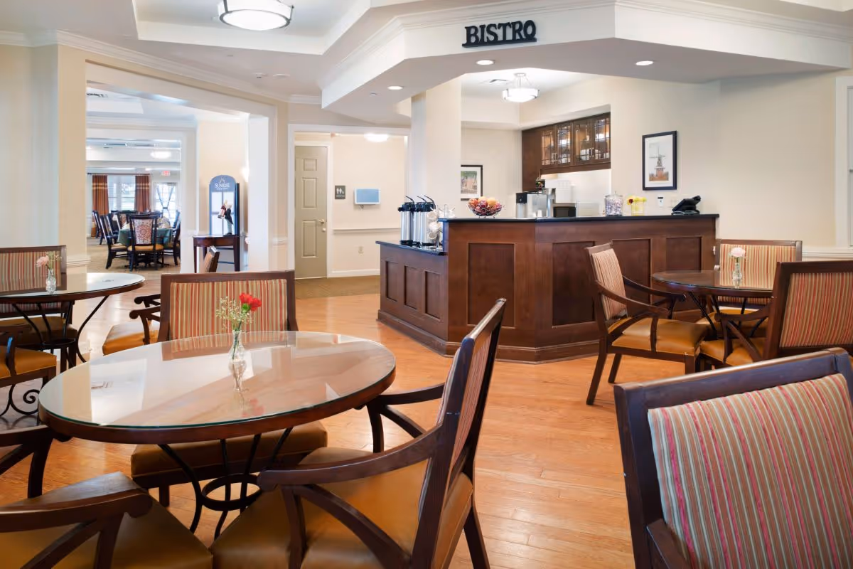 A bright and clean dining area in a senior living facility with round tables and wooden chairs featuring striped upholstery. A wooden counter labeled 'Bistro' is visible in the background with coffee dispensers and a bowl of fruit. The room has hardwood floors, soft lighting, and framed artwork on the walls.