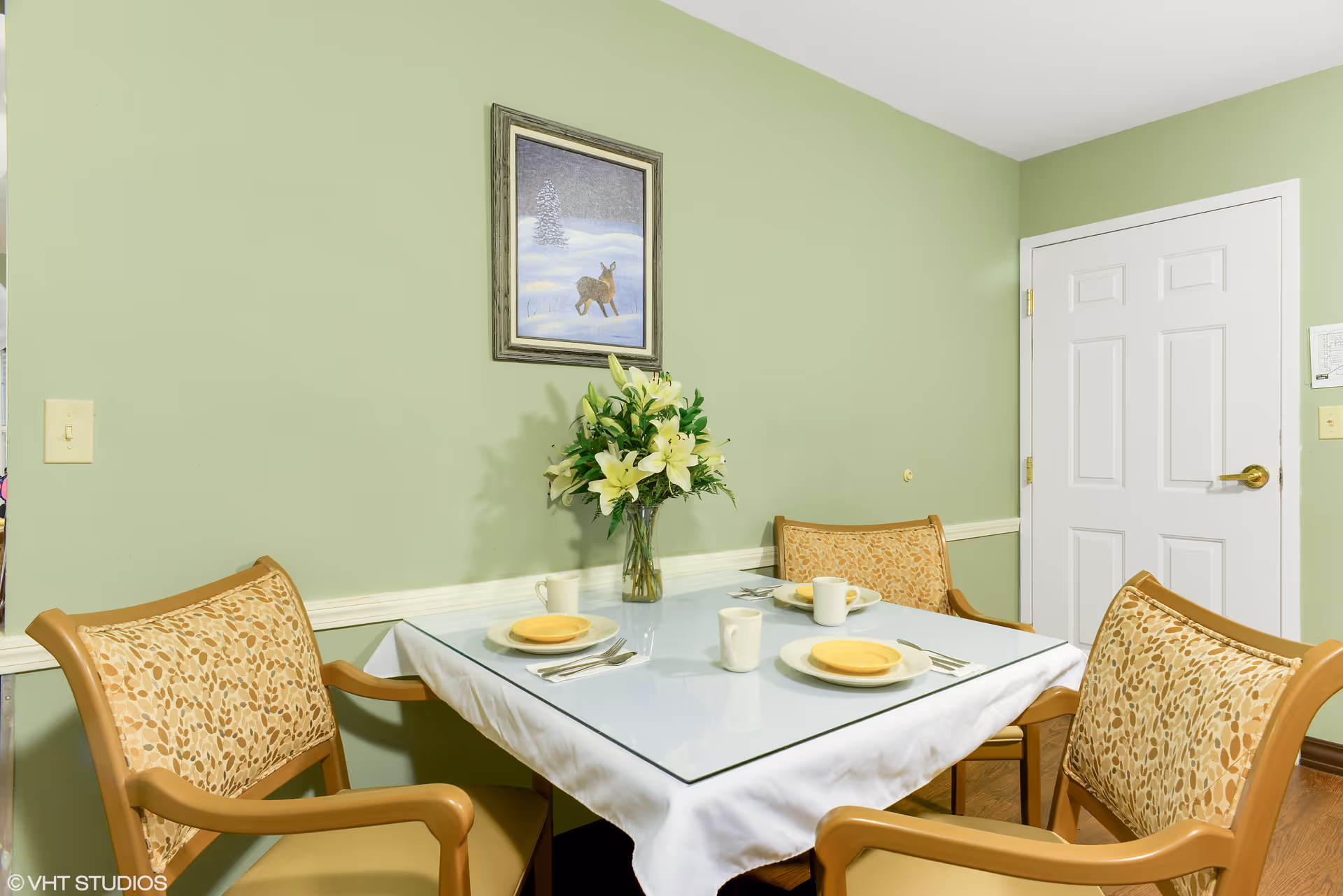 A small dining area with a square table covered with a white tablecloth and glass top. The table is set with four place settings, each with a yellow plate, a white mug, and silverware. There is a vase with white lilies in the center of the table. Four cushioned chairs with patterned upholstery surround the table. The walls are painted light green, and there is a framed picture of a deer in a snowy landscape hanging on the wall. A white door is visible in the background.