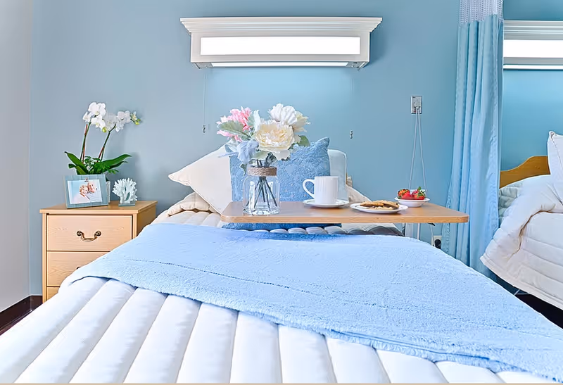 A neatly made bed in a light-blue decorated bedroom with a bedside table, flowers, and a tray holding a mug and plate.