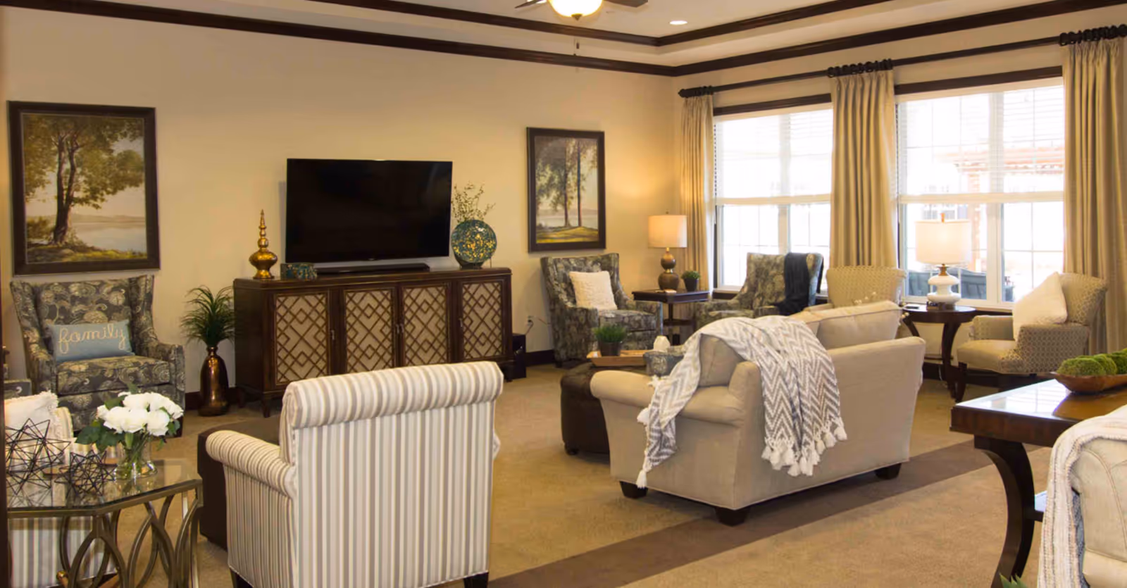 Bright, furnished communal living room with sofas, armchairs, a TV console, and large windows.