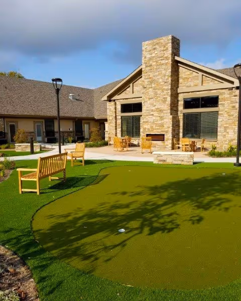 Outdoor area of a senior living facility featuring a putting green with a golf hole, wooden benches, and chairs. The building in the background has a stone facade with large windows and a chimney. The sky is partly cloudy.