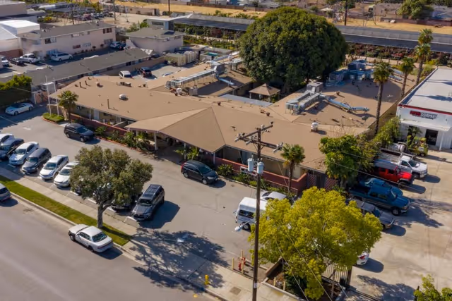 Aerial view of a single-story healthcare building with a covered entrance, adjacent parking lot with parked cars, and surrounding trees.