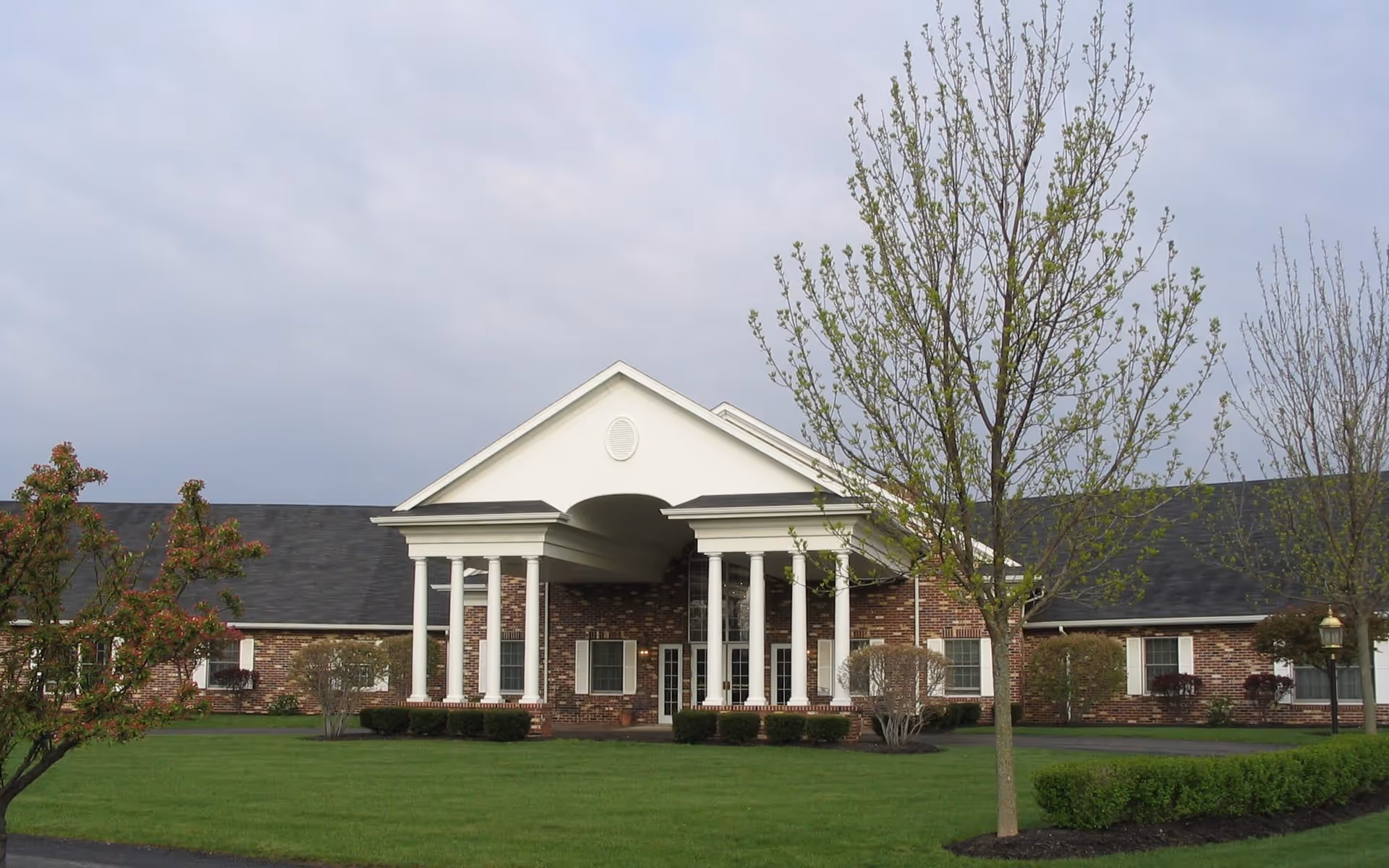 Front exterior view of The Heritage facility showing a brick building with white columns supporting a covered entrance, surrounded by green lawn and young trees under a cloudy sky.