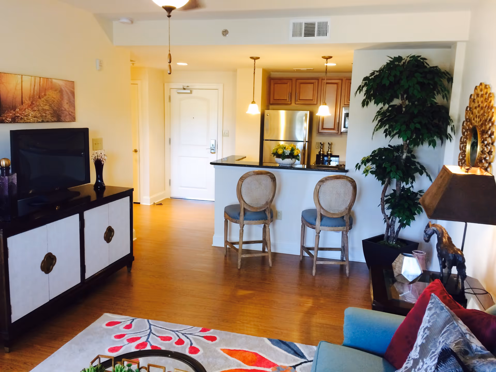 A cozy living room area with a blue armchair adorned with red and gray pillows, a decorative rug with red and orange leaf patterns, a wooden cabinet with a TV on top, and a kitchen area in the background featuring a stainless steel refrigerator, wooden cabinets, and two bar stools at a counter. There is also a tall green plant, a table lamp, and wall decorations including a mirror and a framed picture.