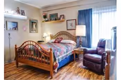 Cozy bedroom with a wooden bed covered by a colorful quilt, nightstand with a lamp, an upholstered recliner by a curtained window, and shelves with framed photos.