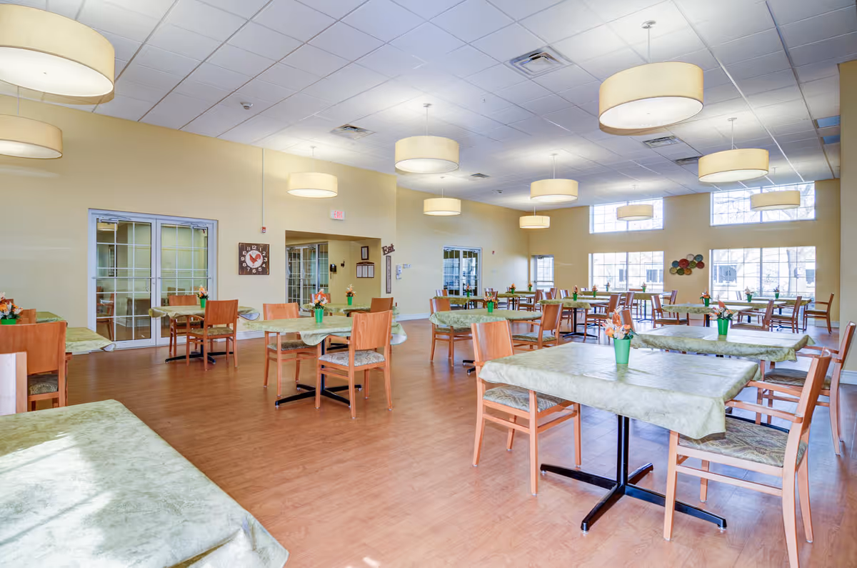 A spacious dining room with multiple tables covered in green tablecloths and wooden chairs. Each table has a small green pot with flowers. The room has large windows letting in natural light, beige walls, and several round ceiling lights. There are double glass doors and a clock on one wall.