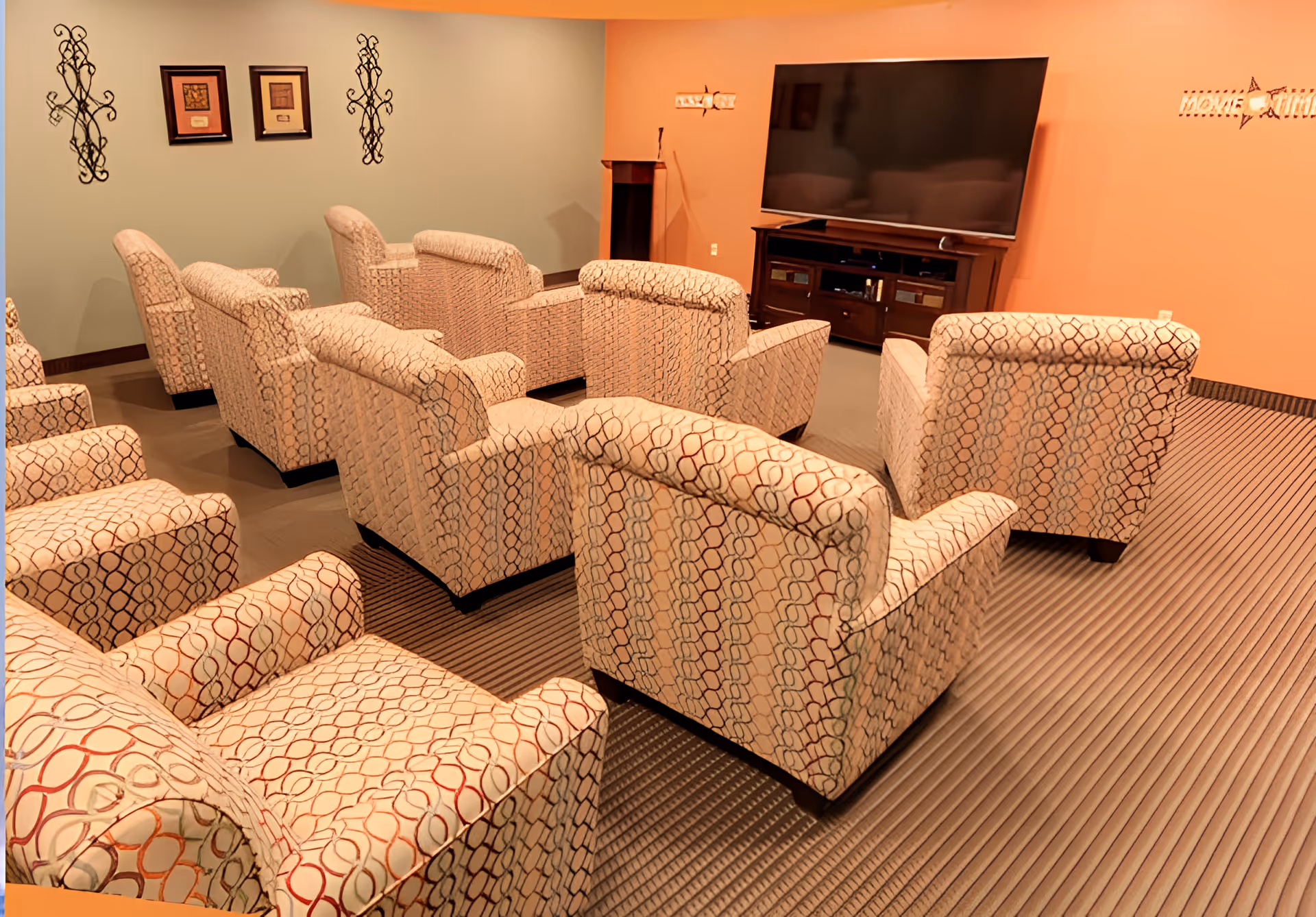 A cozy media room with multiple patterned armchairs arranged in rows facing a large flat-screen TV mounted on a wooden entertainment center. The walls are painted in warm tones of orange and green, decorated with framed artwork and metal wall hangings. The carpet has a striped pattern.