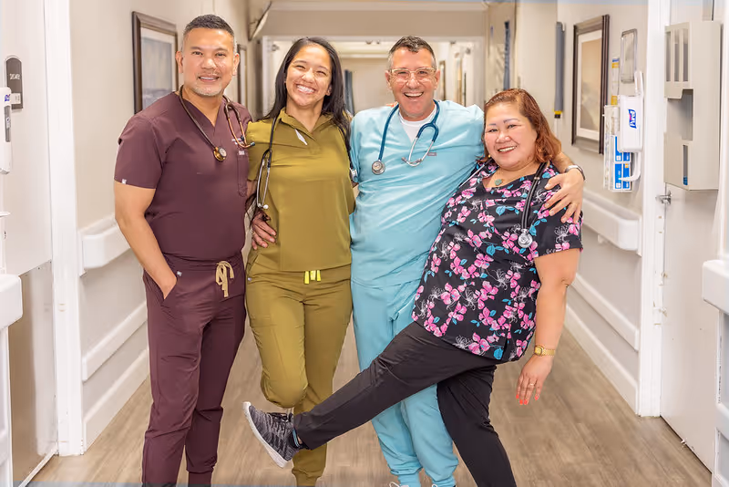 Four healthcare professionals standing together in a hallway, smiling and posing for the camera. They are wearing scrubs in different colors and have stethoscopes around their necks. The hallway has white walls with handrails and framed pictures.