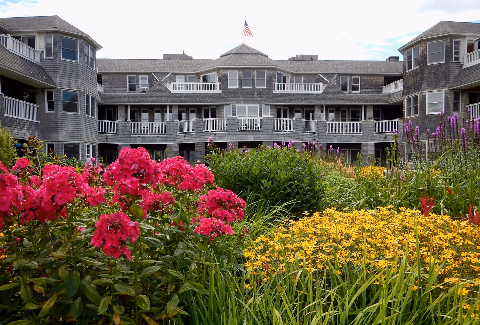 Front of a multi-story gray-shingled retirement building with balconies set behind a colorful garden of pink and yellow flowers.