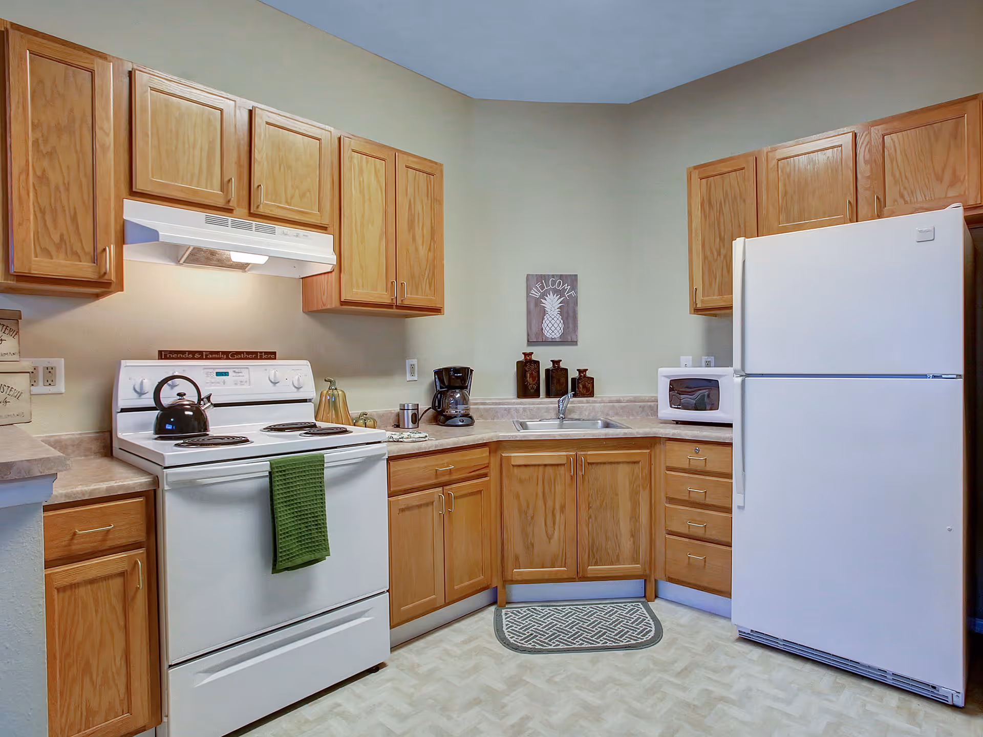 A kitchen with wooden cabinets, a white electric stove with a green towel hanging on the handle, a black kettle on the stove, a coffee maker, a microwave, a white refrigerator, and a small welcome sign with a pineapple on the wall above the sink.