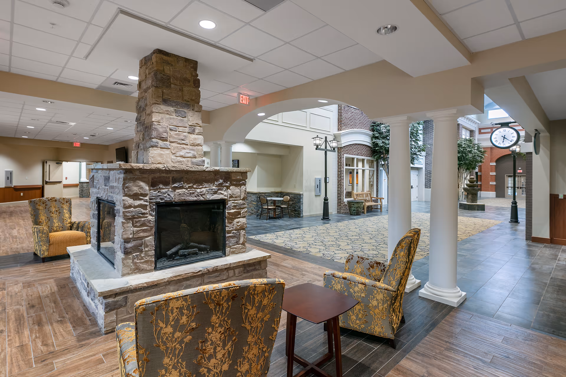 Interior view of a senior living facility featuring a stone fireplace with seating around it, including upholstered armchairs and a small wooden table. The space has a mix of wood and tile flooring, white columns, and an open area with a patterned carpet. There is a large clock on a lamppost and some greenery in the background, creating a welcoming and comfortable common area.