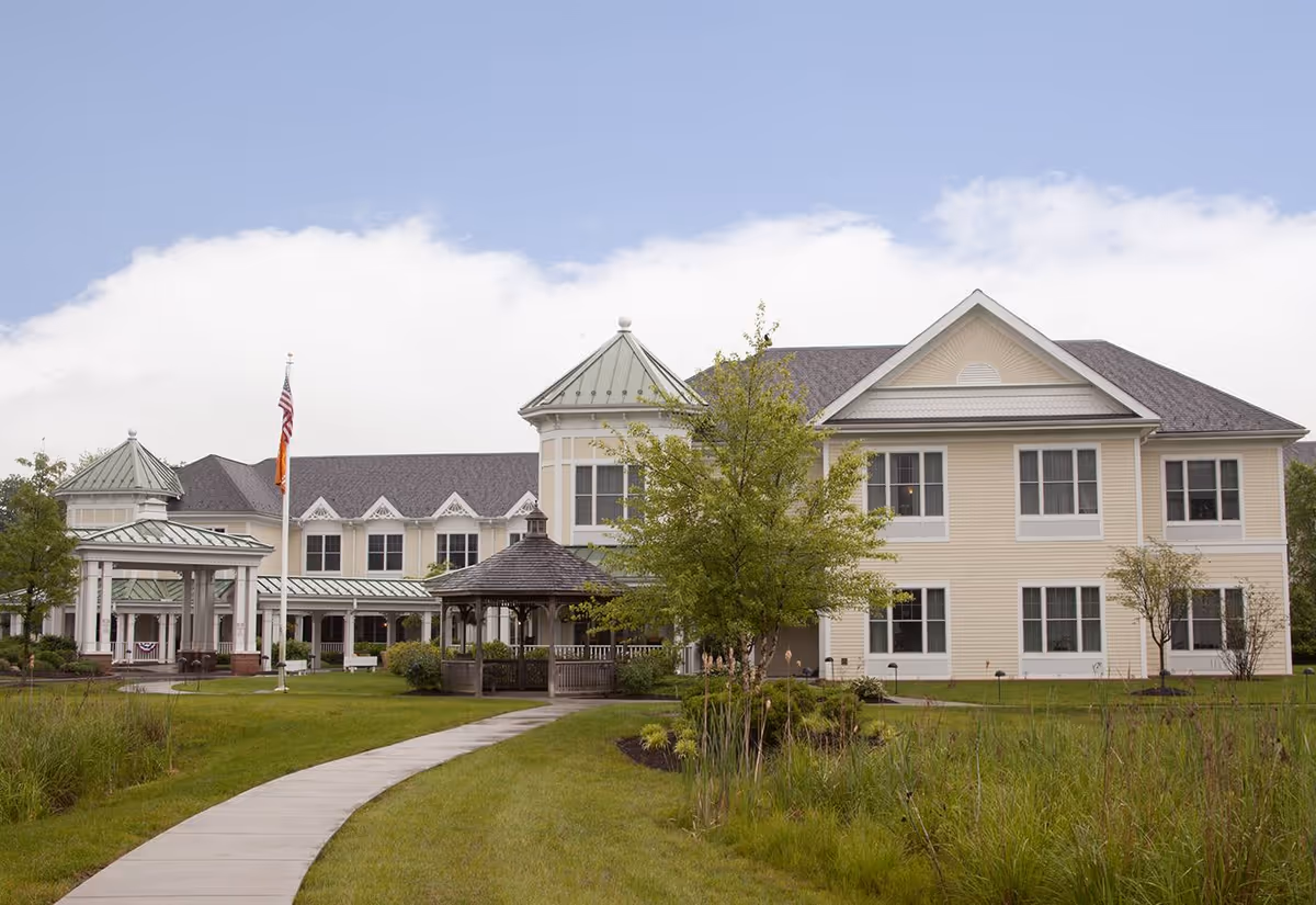 Exterior view of a two-story senior living facility building with beige siding and green metal roofs on some sections. A paved walkway curves through a well-maintained lawn with trees and shrubs leading to a gazebo and the building entrance. An American flag is flying on a flagpole near the entrance under a partly cloudy sky.