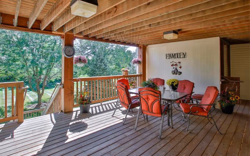 Covered wooden deck with a glass-top table and six red cushioned chairs. The deck overlooks a green outdoor area with trees. There are hanging flower pots and potted plants around the deck. A wall decoration with the word 'FAMILY' and floral designs is mounted on the wall.