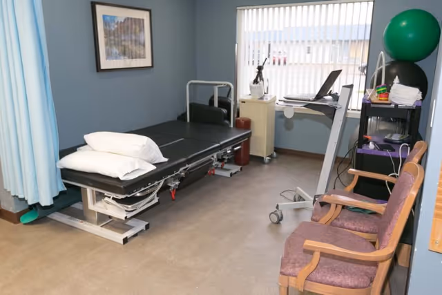 Small rehabilitation therapy room with an adjustable treatment table, exercise equipment, chairs, and a window with vertical blinds.
