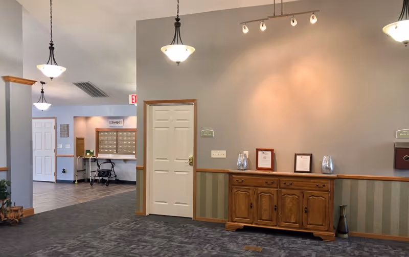 Interior view of a senior living facility hallway with a wooden cabinet against a gray wall, decorative lighting fixtures hanging from the ceiling, and a section with mailboxes and a walker in the background.