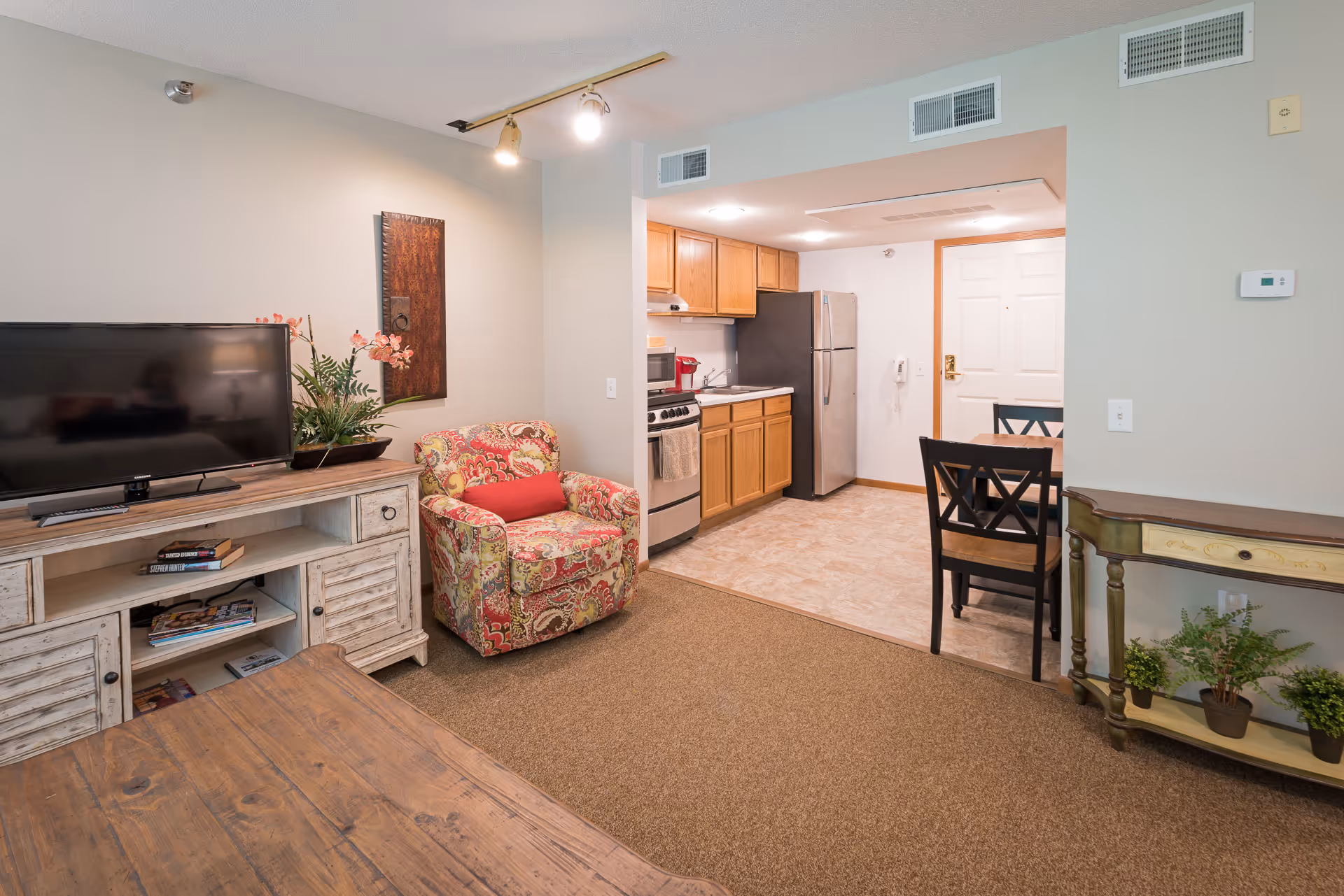 Interior view of a senior living facility apartment showing a cozy living area with a floral armchair, a wooden TV stand with a flat-screen TV, and a small kitchen with wooden cabinets, a stove, and a refrigerator. A dining table with two chairs is positioned near the entrance door. The room has a mix of carpet and tile flooring and neutral-colored walls.