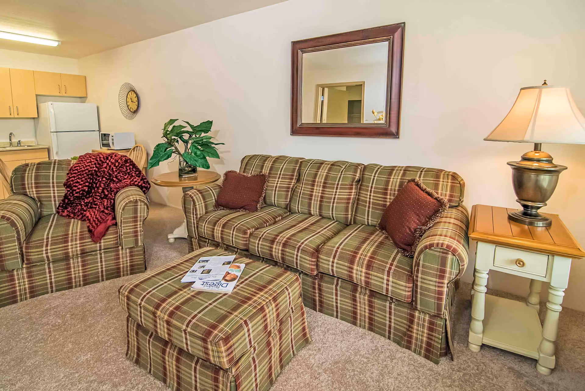 Plaid sofa, matching armchair and ottoman in a living room with a side table and lamp, mirror on the wall, and a small kitchenette visible in the background.