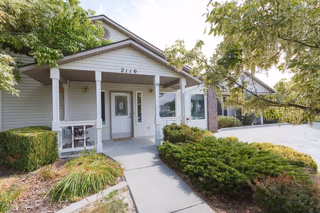 Front exterior view of a single-story building with a covered porch entrance, white siding, and the number 2110 above the door. There are shrubs and trees surrounding the concrete walkway leading to the entrance.