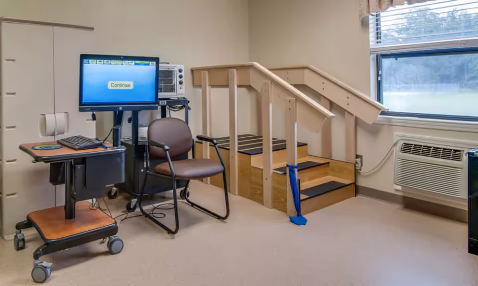 Small therapy room with a computer on a rolling cart, a chair, a short wooden practice stair with handrails, and a window with an air conditioner.