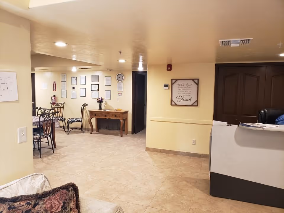 Interior view of a senior living facility showing a reception desk on the right, a small table with a flower vase against the wall, several chairs around a table on the left, and framed certificates and a clock on the far wall. The walls are painted light yellow and the floor is tiled.