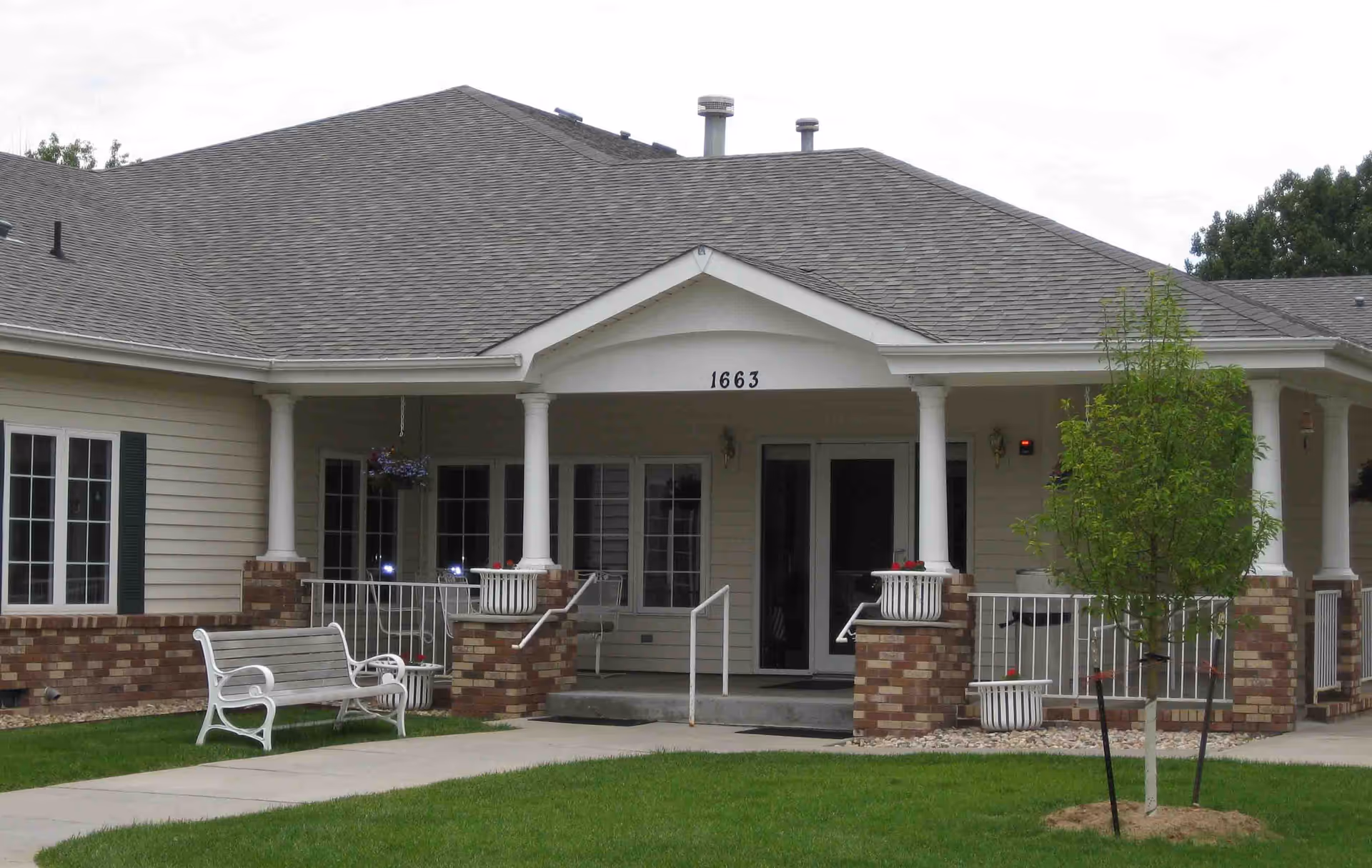 Front exterior view of a single-story building with a gray shingled roof, beige siding, and brick accents. The entrance has white columns supporting a small portico with the number 1663 above the door. There is a white bench on the left side of the walkway leading to the entrance, and a small tree planted on the right side in a grassy area.
