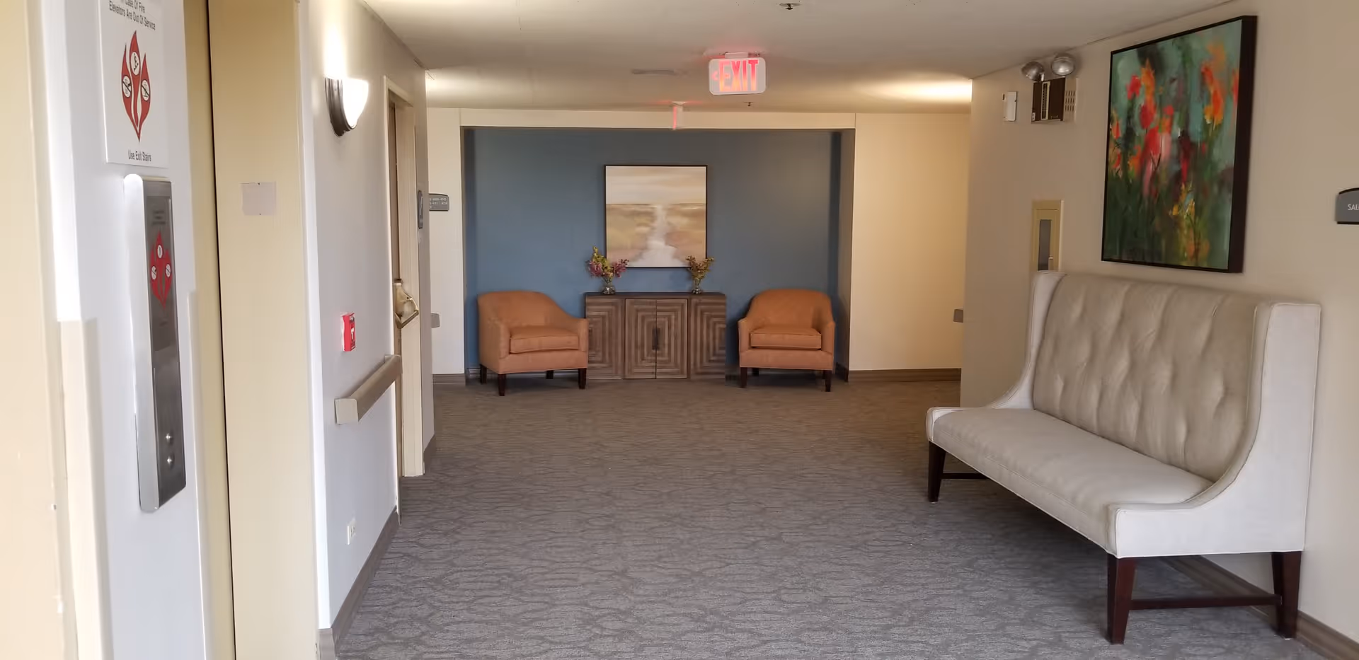 A hallway in a senior living facility with an elevator on the left, two orange armchairs and a wooden cabinet against a blue accent wall at the end, and a white cushioned bench on the right wall beneath a colorful abstract painting. The carpet is patterned and the walls are light-colored with soft lighting fixtures.