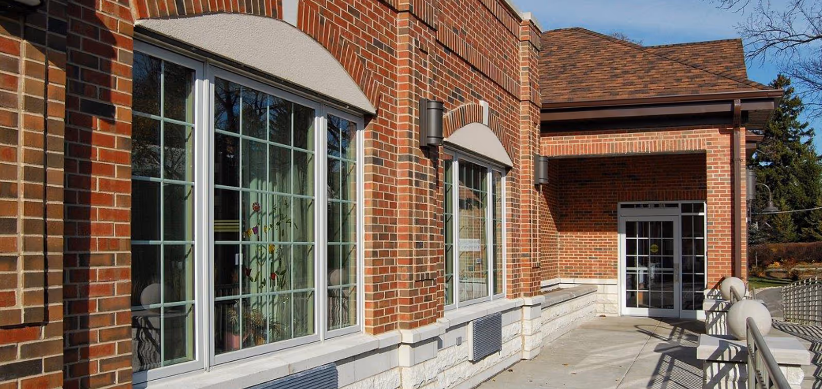 Brick building facade with large multi-pane windows and a covered entrance along a concrete walkway.