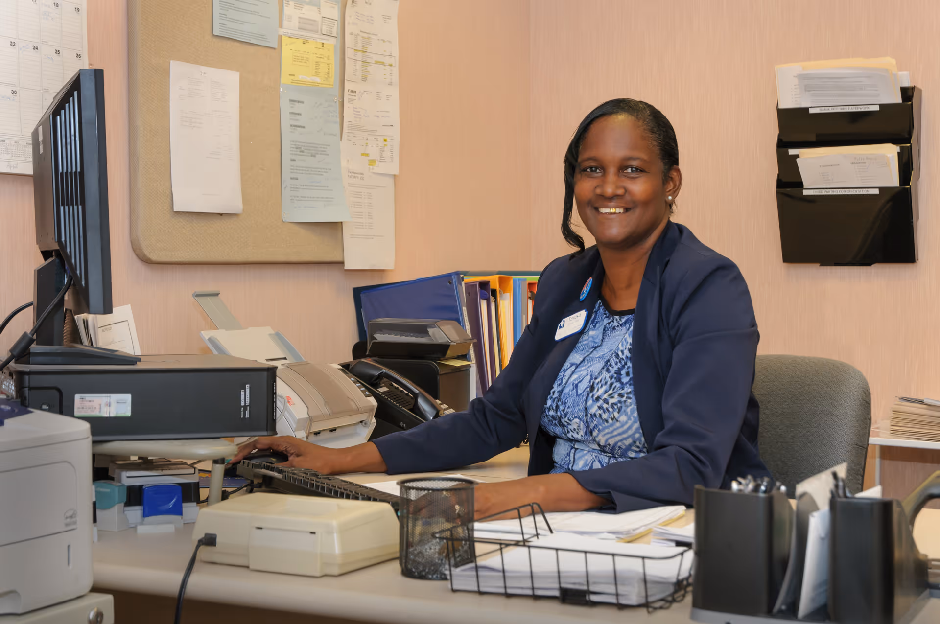 A woman wearing a blue blazer and patterned blouse is sitting at a desk in an office environment, smiling at the camera. The desk has office equipment including a computer, printer, telephone, and various papers and office supplies. Behind her, there is a bulletin board with pinned papers and a wall-mounted organizer with documents.
